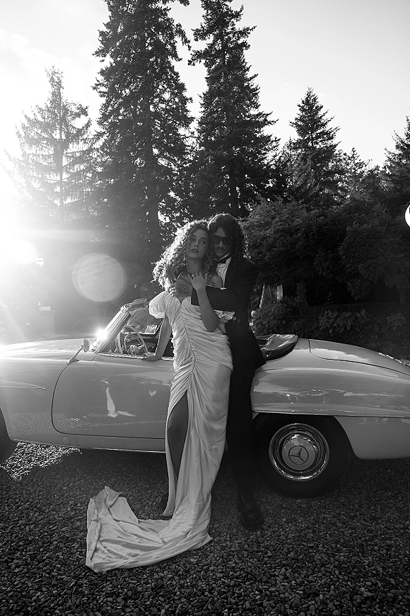 Couple portrait of bride and groom by car, groom hugging bride in tuxedo and sunglasses beside a vintage convertible in sun flare