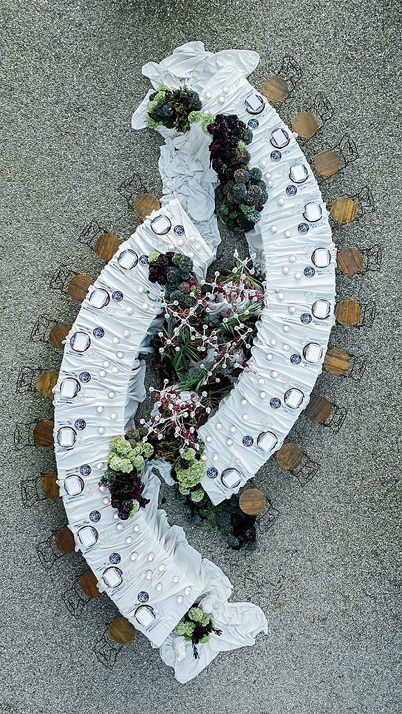 Reception table layout with curved banquet tables in white linens, candlelit place settings, and floral greenery centerpieces on gravel