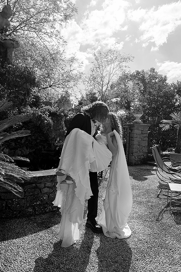 Wedding kiss portrait of groom carrying bride in a dip, her bridal veil flowing, in a gravel courtyard by a stone wall and trees