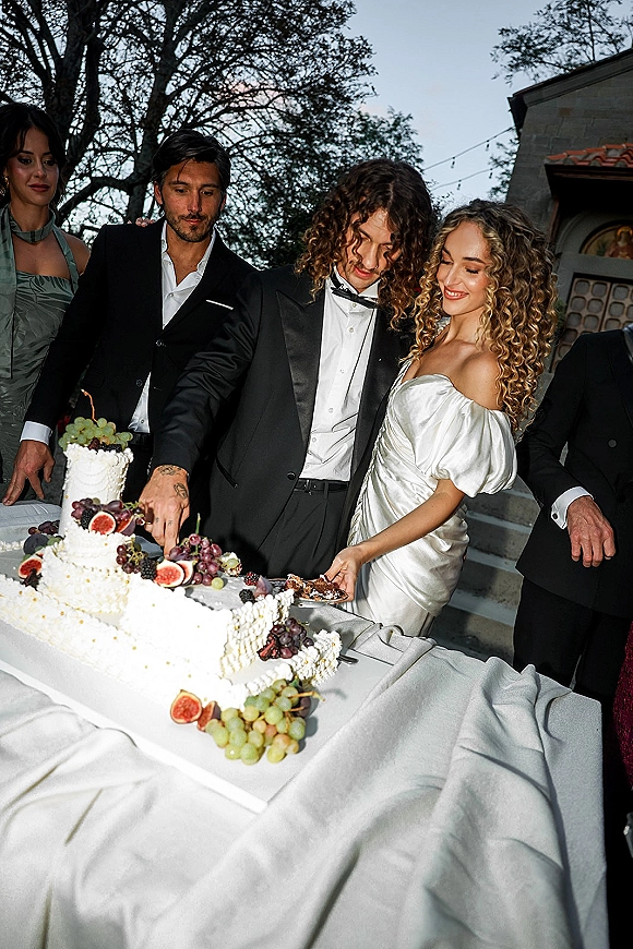 Wedding cake cutting as bride in an off-shoulder satin dress and groom in black tux slice a fruit-topped cake under string lights outdoors