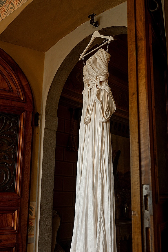 Wedding dress hanging on a hanger from a wall hook, strapless wedding dress draped in an arched doorway with wooden door and stone arch