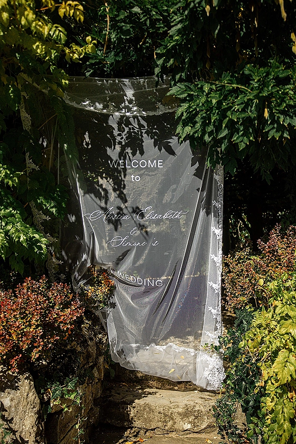 Wedding welcome sign, acrylic wedding welcome sign with white lettering and lace trim, draped in tulle on stone steps amid garden foliage