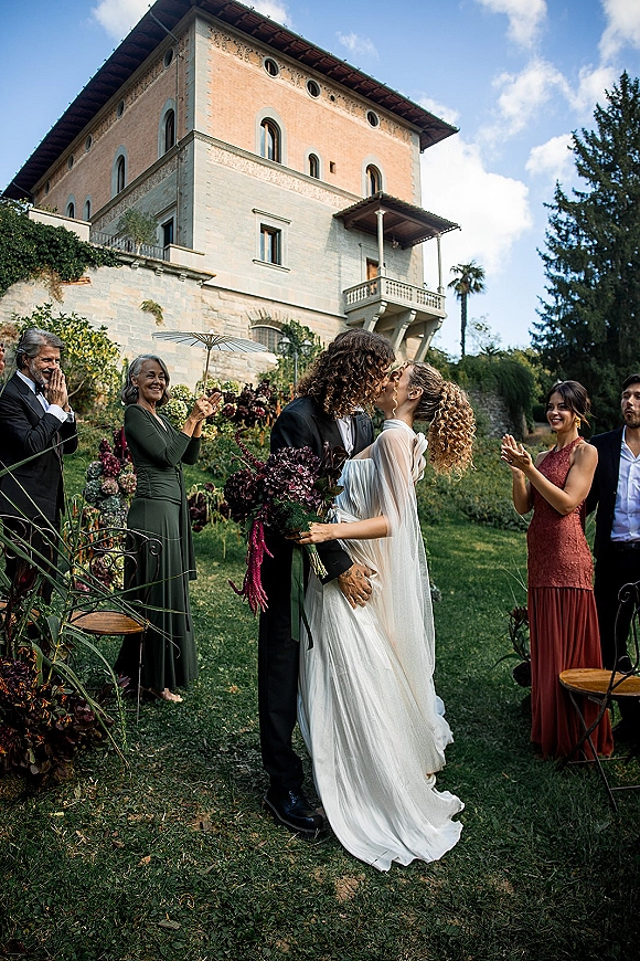 Wedding kiss as the bride in a veil holds a bouquet and the groom in a black suit kisses her on a villa lawn with cheering guests