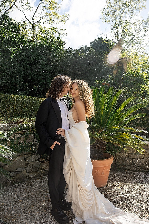 Couple portrait of bride and groom embrace as he kisses her forehead, her draped gown and shawl glowing in sunlit garden by stone wall