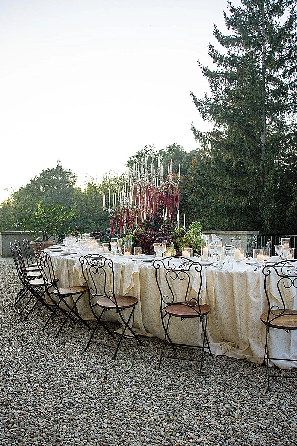 Reception tablescape with round table settings, neutral linens, glassware, taper candles and candelabra centerpiece on a stone terrace outdoors