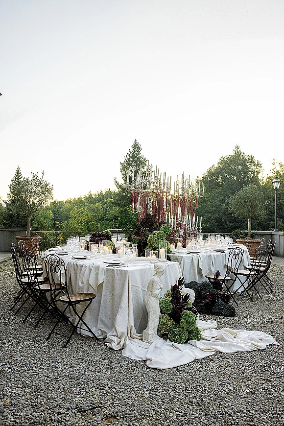 Reception tablescape with taper candles, burgundy ribbons, and florals on round tables with black wrought iron chairs on a gravel patio