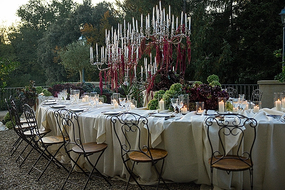 Reception tablescape with ivory linens on a long banquet table, silver candelabras and taper candles, floral centerpieces on a garden terrace