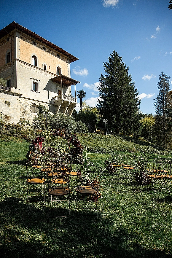 Outdoor ceremony setup with garden wedding ceremony seating, wrought iron chairs, and burgundy florals on a lawn before a stone villa backdrop