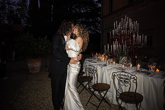 Wedding kiss as the bride and groom kissing beside a long banquet table with candlelit candelabra in a courtyard at night