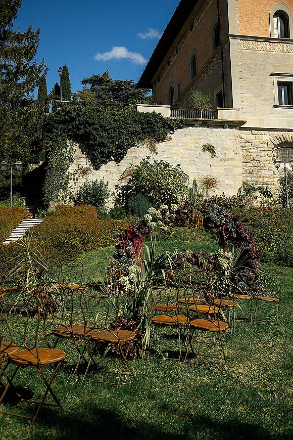 Ceremony setup with outdoor garden ceremony chairs facing a floral ceremony arch, aisle florals on a lawn by an ivy stone wall under blue sky