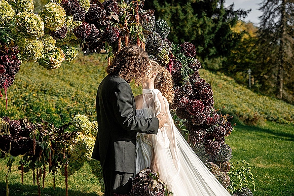 Wedding kiss during a ceremony kiss moment beneath an asymmetrical hydrangea and greenery floral arch on a grassy hillside outdoors
