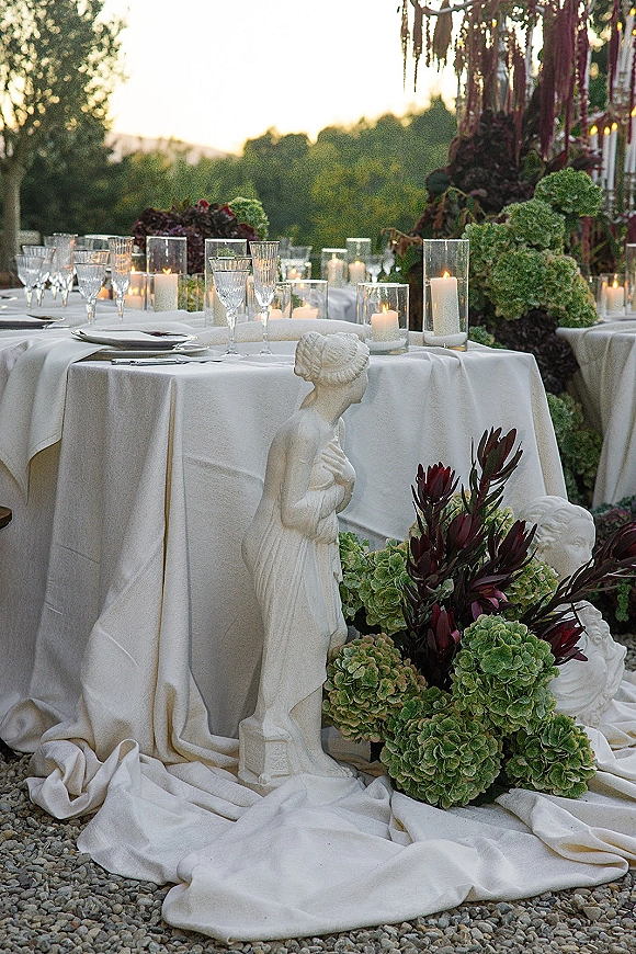Reception tablescape with white linen table set with glass hurricane candles, hydrangea and burgundy florals, statues at sunset outdoors