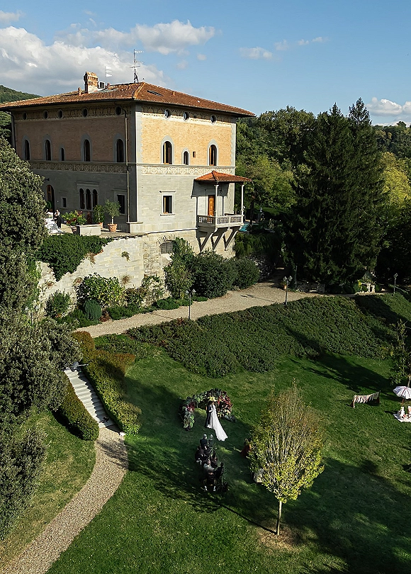 Outdoor wedding ceremony with bride at a floral arch, chairs on garden lawn by a stone villa under blue sky clouds