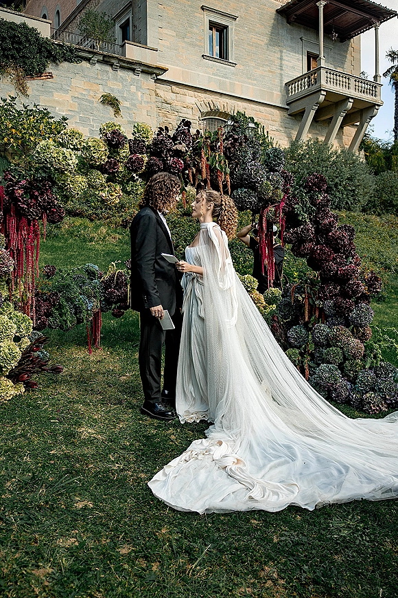 Wedding vows as bride in a cape and long train reads from vow book facing groom in tux under hydrangea floral arch at stone villa lawn ceremony