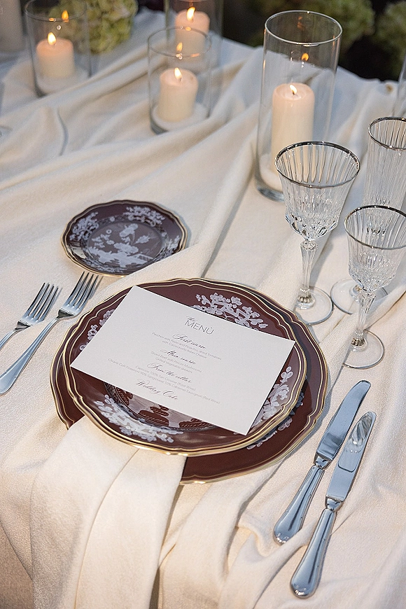 Reception tablescape with wedding place setting, menu card on burgundy patterned plates, crystal glasses, and votive candles before blurred florals