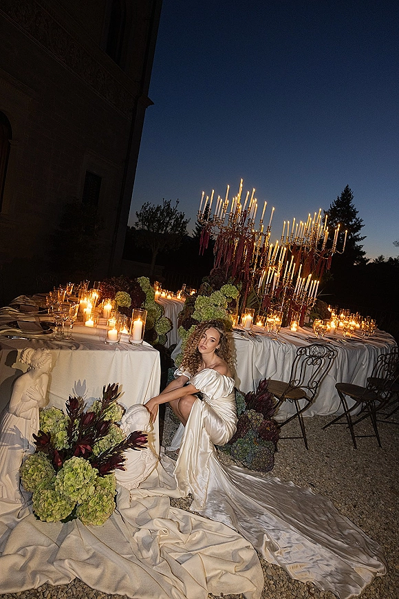 Bridal portrait of a bride in an off the shoulder wedding dress with a long satin train, seated by candlelit candelabras on a twilight terrace