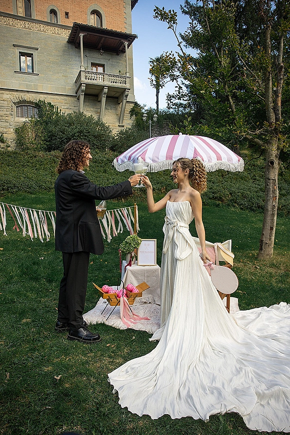 Wedding toast as bride and groom clink champagne coupe glasses on a garden lawn with striped parasol and stone building backdrop