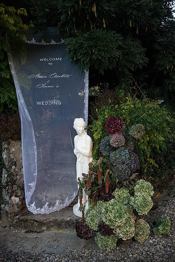 Wedding welcome sign on a clear acrylic welcome sign with calligraphy, draped lace veil and hydrangeas by stone steps in a garden path