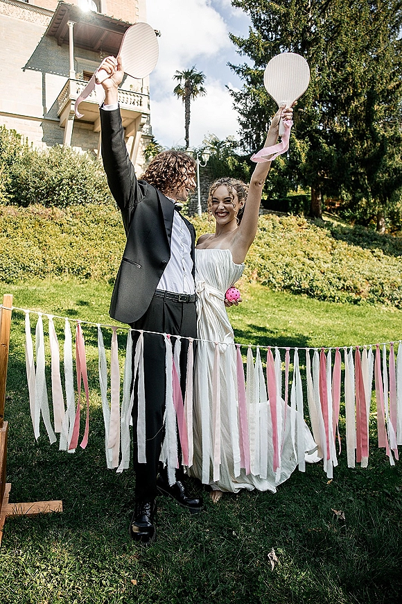 Couple portrait of bride and groom playing wedding lawn games, cheering with paddles by a badminton net on a villa garden lawn with pink ribbons