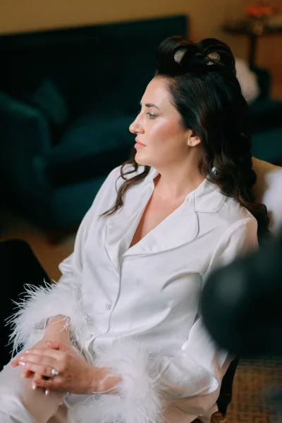 Bridal getting ready in white satin pajamas with feather trim cuffs, showing engagement ring and manicure while seated indoors on a sofa