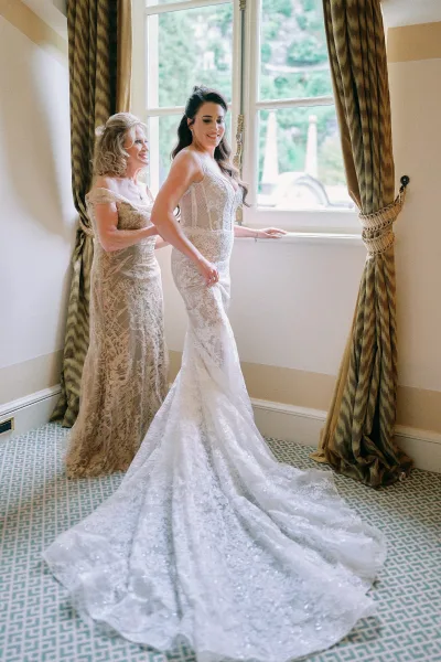 Bride getting ready as mother helps her into a lace mermaid wedding dress by a large window, long train trailing on patterned carpet