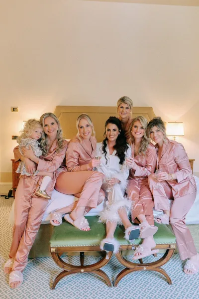 Bridesmaids getting ready in bridal party pajamas, holding champagne glasses on a bed in a hotel bedroom with lamps and headboard