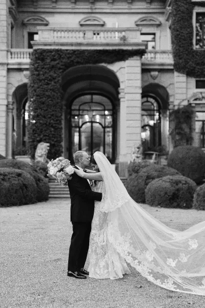 Wedding couple portrait in black and white, groom kissing bride’s forehead as she holds a bouquet, long lace veil trailing in ivy courtyard