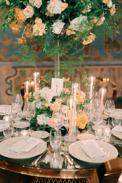 Reception tablescape with wedding table centerpiece of tall roses and greenery in a clear vase, taper candles on a mirrored table, soft lights behind