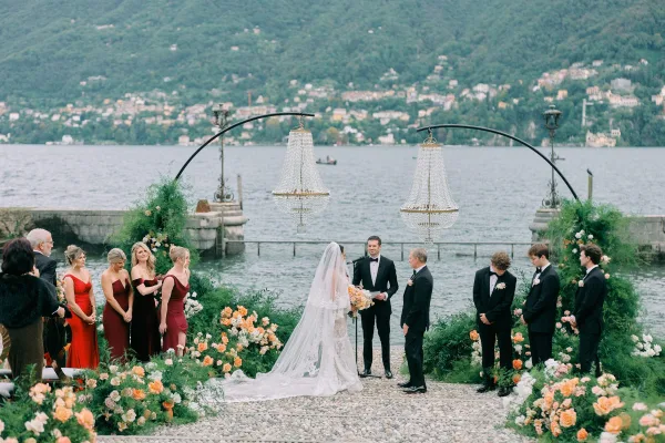 Wedding ceremony with bride and groom at the altar under a chandelier greenery arch, overlooking a mountain lake on a stone terrace