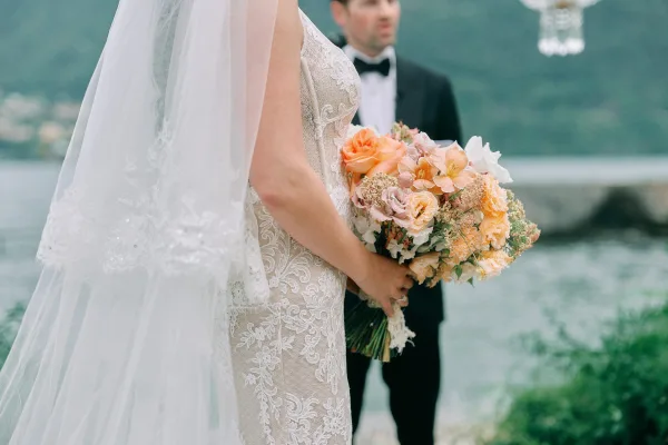 Bride holding bouquet with peach and blush bouquet in lace wedding dress and veil, groom in black tuxedo by a lakeside with mountains