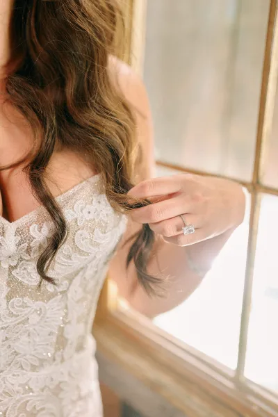 Bridal portrait with engagement ring close up as the bride touches her loose curls in warm window light beside paneled windows