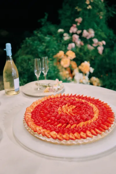 Wedding cake on a wedding dessert table topped with sliced strawberries and raspberries beside champagne flutes in a garden at night