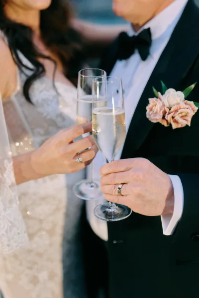 Wedding toast as bride and groom clink champagne flutes, sparkling wine catching light with rings visible against blurred outdoors