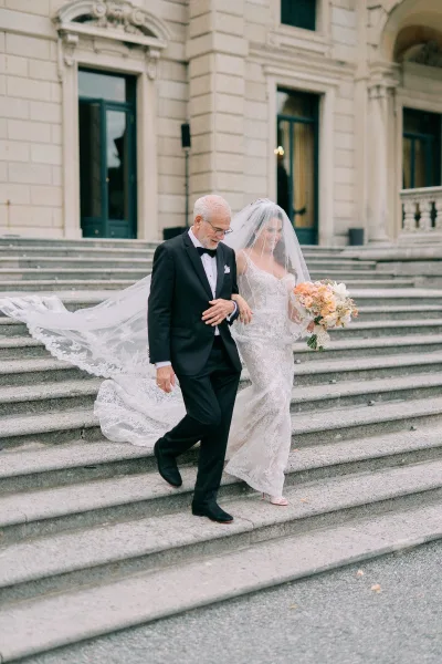 Wedding processional as bride walking with father down stone steps, cathedral veil trailing, peach bouquet in hand before grand doorway