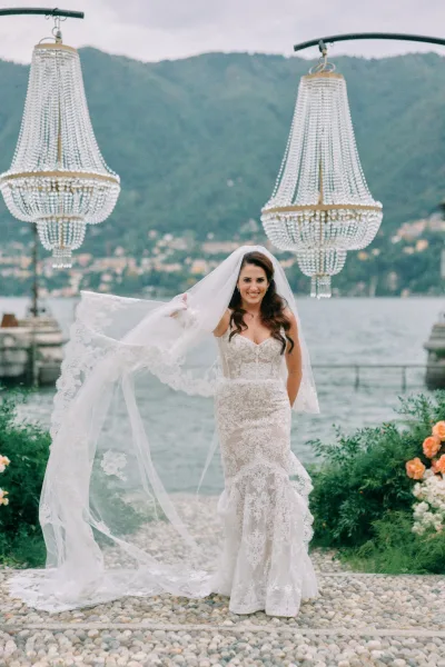Bridal portrait of a bride holding veil in a lace mermaid wedding dress on a stone path with chandeliers, lake and mountains behind
