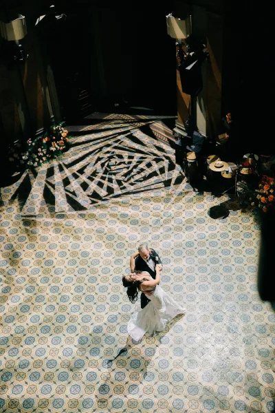 First dance as bride and groom dip for a kiss on a patterned tile floor, sunlit shadows in an indoor hall with band and florals