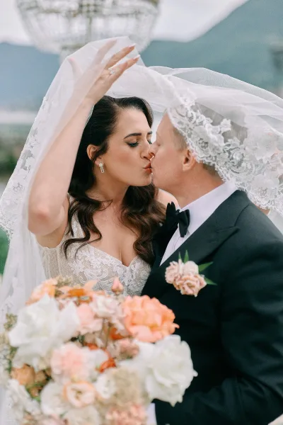 Wedding kiss portrait of bride and groom kissing under a lace veil, bride holding a peach-and-white rose bouquet outdoors amid greenery under overcast sky