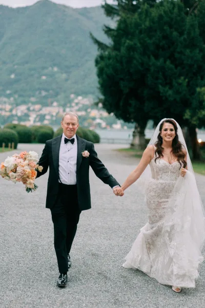 Bride and groom walking hand in hand down a gravel path, bride’s long veil flowing, with mountains and lake in the background