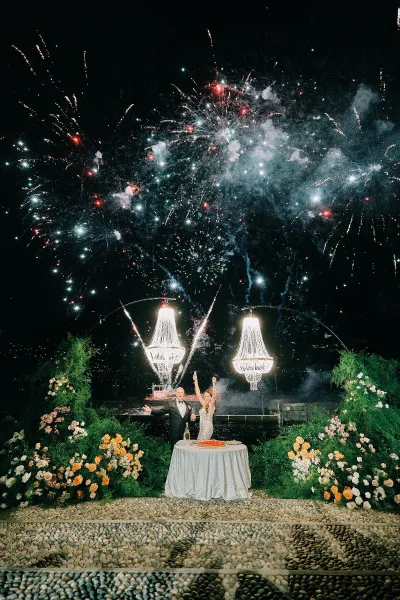 Wedding fireworks moment as newlyweds in tuxedo and wedding dress stand by a cake table on a stone terrace by the waterfront at night