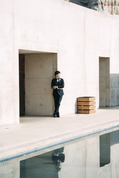 Groom portrait in a black tuxedo with boutonniere and glasses, leaning by a concrete wall near a reflecting pool at a modern venue exterior