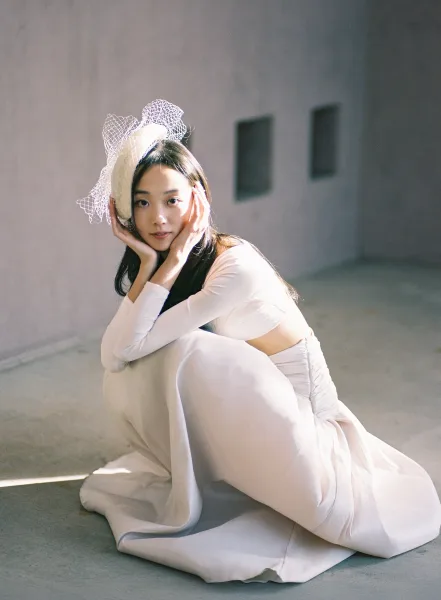 Bridal portrait of a bride in a fitted wedding dress with a birdcage veil, posed in window light against a white wall with frames