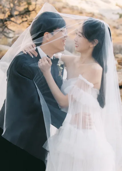 Couple portrait of bride and groom under veil, smiling as she adjusts his bow tie; he wears a black tuxedo in a rocky treed landscape