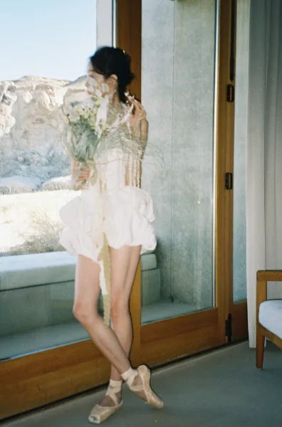 Bridal portrait of a bride holding bouquet in a short wedding dress by a floor-to-ceiling window with ocean view coastline beyond
