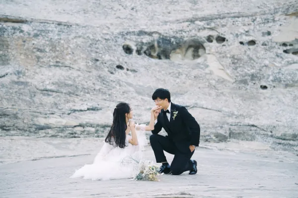 Couple portrait of bride and groom kneeling as he kisses her hand beside a rocky shoreline, her dress train trailing on sand