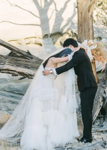 Wedding kiss as bride and groom embrace, her cathedral veil flowing over strapless gown by a rock wall with desert shadows
