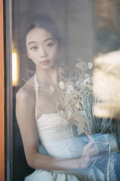 Bridal portrait of bride holding bouquet in a strapless wedding dress by a window, soft reflections on glass and wooden frame in gentle light