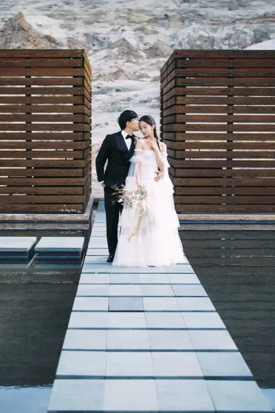 Couple portrait of groom kissing bride’s forehead, her veil and white bouquet flowing beside a reflecting pool and wood slat wall backdrop