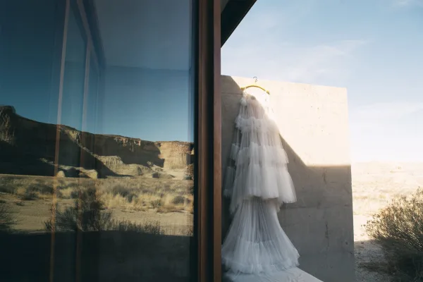 Wedding dress hanging on a hanger, tiered tulle ruffle skirt catching sunlight against a concrete wall with desert mountains beyond