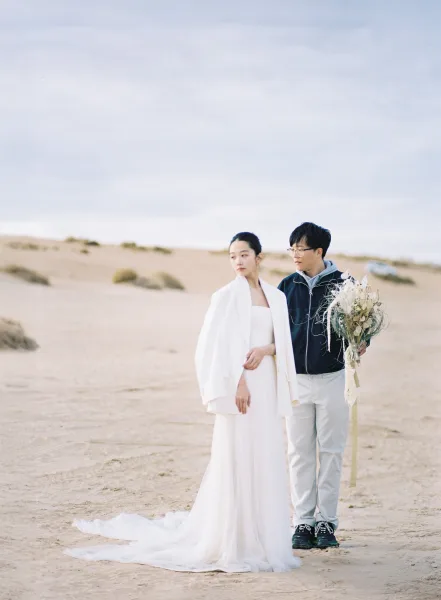 Couple portrait in desert engagement photos, bride in strapless gown with long train and groom in white blazer on sand dunes holding bouquet ribbon