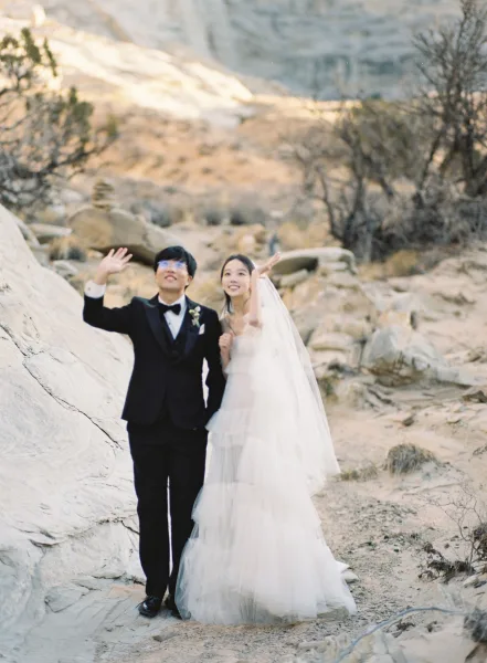 Couple portrait of bride and groom waving, her long veil flowing beside his tuxedo, in a rocky desert landscape with boulders